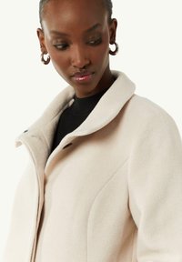 Hellbeige Wollmantel mit strukturiertem Kragen, schlanken Knöpfen und seitlichen Nähten, kombiniert mit einem schwarzen gerippten Rollkragenpullover.