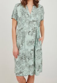 Mint-vihreä mekko, jossa on lyhyet hihat, V-pääntie, musta kukkakuosi, laskokset edessä ja sivutaskut. Sujuva kankaan pinta.