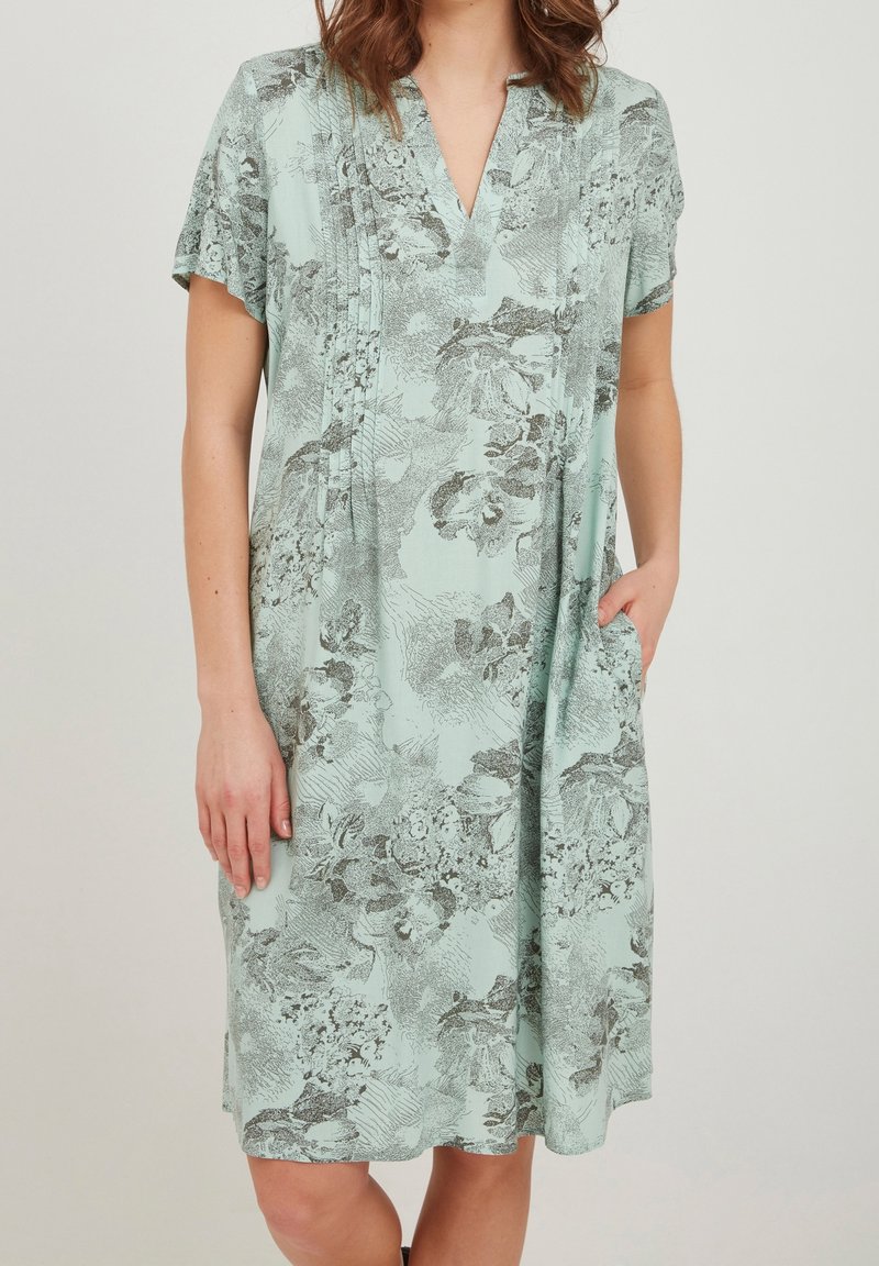 Mint-vihreä mekko, jossa on lyhyet hihat, V-pääntie, musta kukkakuosi, laskokset edessä ja sivutaskut. Sujuva kankaan pinta.