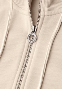 Sweat à capuche beige zippé avec un tissu texturé, des ourlets côtelés et un tirette de zip argenté avec logo. Gros plan sur la fermeture éclair et les détails de couture.