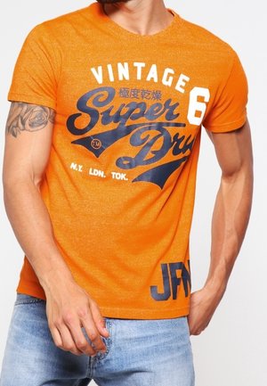 Mężczyzna w jasnopomarańczowej, vintage koszulce Super Dry z czarno-białym napisem, w jasnoniebieskich dżinsach, pokazujący tatuaż na lewym ramieniu.