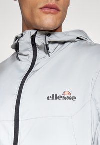 Hellgrauer Windbreaker mit schwarzem Reißverschluss, verstellbarem Kordelzug in der Kapuze und "ellesse"-Logo in Schwarz und Rot. Glatte Textur, athletisches Design.