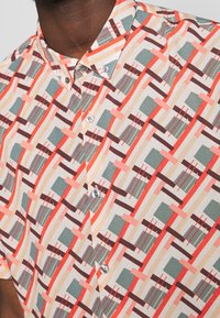 Camicia a maniche corte con bottoni caratterizzata da un motivo geometrico in corallo, marrone, teal e beige su uno sfondo chiaro. Tessuto in cotone, colletto classico.