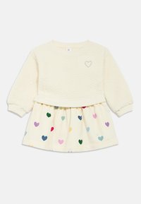 Krém színű pulóver texturált puha anyagú törzzsel, szív hímzéssel és krém szoknyával, amelyet különböző méretű, színes szívminták díszítenek.