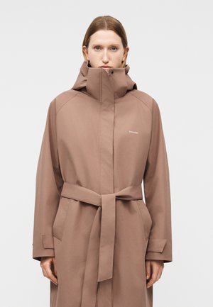Γυναίκα που φοράει ένα ανοιχτό καφέ παλτό τρανς στυλ με ζώνη, ψηλό γιακά και κουκούλα, στέκεται μπροστά σε ένα απλό φωτεινό φόντο.