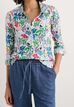 Frau trägt ein weißes Hemd mit bunten Blumenmustern und hochgekrempelten Ärmeln, kombiniert mit blauen Jeanshosen mit Kordelzug.