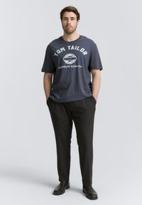 Szary t-shirt z napisem "TOM TAILOR" i "PREMIUM QUALITY", połączony z ciemnymi dopasowanymi spodniami i czarnymi butami. Prosty, luźny krój.