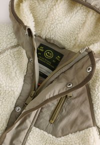 Veste en polaire crème texturée avec un extérieur en nylon beige. Dotée d'une double fermeture éclair, de boutons-pression et d'une étiquette intérieure avec un visage souriant.