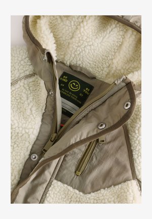 Tekstureret cremefarvet fleecejakke med beige nylonyderside. Har dobbelte lynlåse, trykknaplukninger og en indvendig etiket med et smilende ansigt.