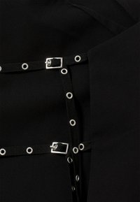 Tissu noir avec des éléments métalliques, comprenant des sangles passant à travers des œillets circulaires et un détail de boucle rectangulaire.