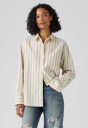 Femme aux cheveux bouclés portant une chemise ample à rayures jaunes et violettes et un jean bleu déchiré, debout avec une main dans la poche.