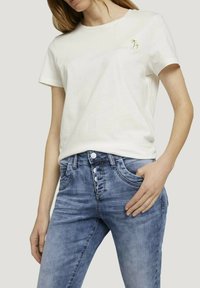 Vit kortärmad t-shirt med en liten palmtree-brodyr på bröstet, parat med ljusblå denimjeans med fem knappar och framfickor.