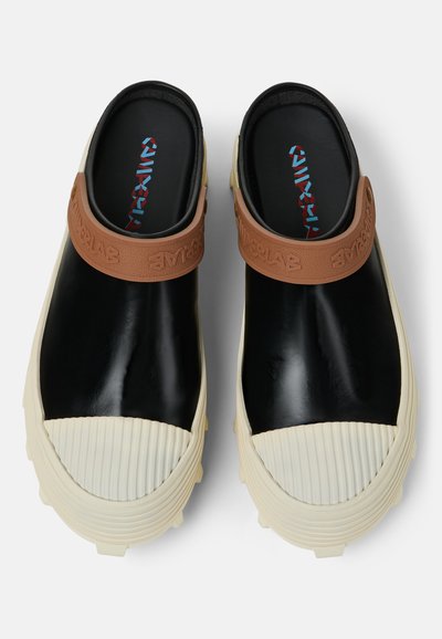 Svarta gummiklackar med en bred vit ribbad tå, beige texturerad rem och mönstrade innersulor. Har en slät, glänsande ovandel och texturerad sula.