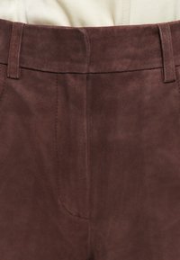 Nærbillede af en mørkebrun ruskindsbuks talje med bæltestropper og syningsdetaljer på beige baggrund.