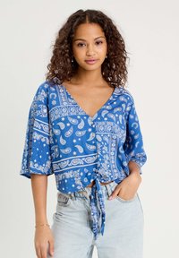 Blouse bleue à manches courtes avec un imprimé paisley, décolleté en V et détail de nouage à l'avant. Fabriquée en tissu léger, elle se marie bien avec un denim clair.
