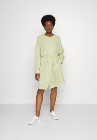 Vestido camiseiro verde-claro, com botões e cinto na cintura, mangas compridas e bolsos laterais. Usado com ténis brancos. Textura suave.