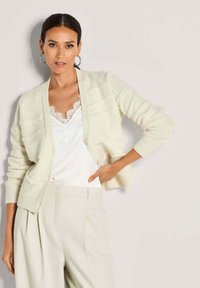 Frau trägt eine crèmefarbene Strickjacke über einem weißen Satinoberteil mit Spitze und hoch geschnittene, gefaltete Hosen, posiert mit der Hand auf der Hüfte vor einem einfarbigen Hintergrund.