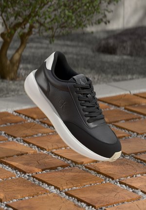 Sneaker in pelle nera con finitura testurizzata, suola bianca e lacci neri. Presenta un logo argento sulla parte superiore e un design elegante.