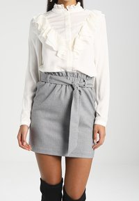 Bílá blůza s volánkovým límcem a přední částí, v kombinaci se šedou kostkovanou minisukní s vázáním v pase a detailem opasku.