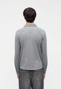 Γκρι μακρυμάνικο πόλο πουκάμισο με γιακά που διπλώνει και μανσέτες με κουμπιά, κατασκευασμένο από μαλακό ύφασμα, φορεμένο με σκούρα γκρι τζιν.