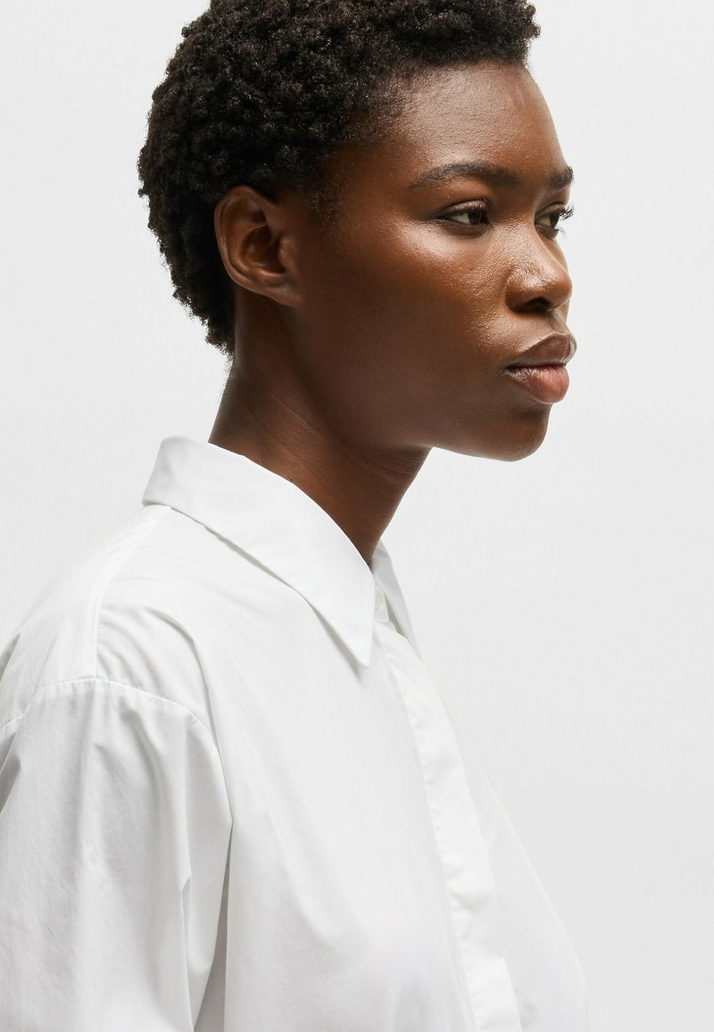 Camisa blanca de botones con cuello clásico, con una textura suave y un corte holgado, mostrando un perfil lateral sobre un fondo neutro.