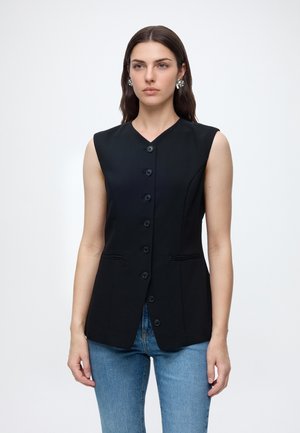 Femme portant un gilet noir sans manches boutonné avec des poches latérales et un jean bleu, debout devant un fond clair uni.