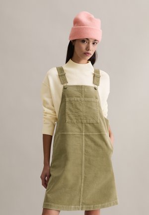 Sukienka na szelkach z oliwkowego sztruksu z regulowanymi paskami, dużą kieszenią z przodu, noszona na kremowym swetrze z dzianiny, uzupełniona różową czapką beanie.