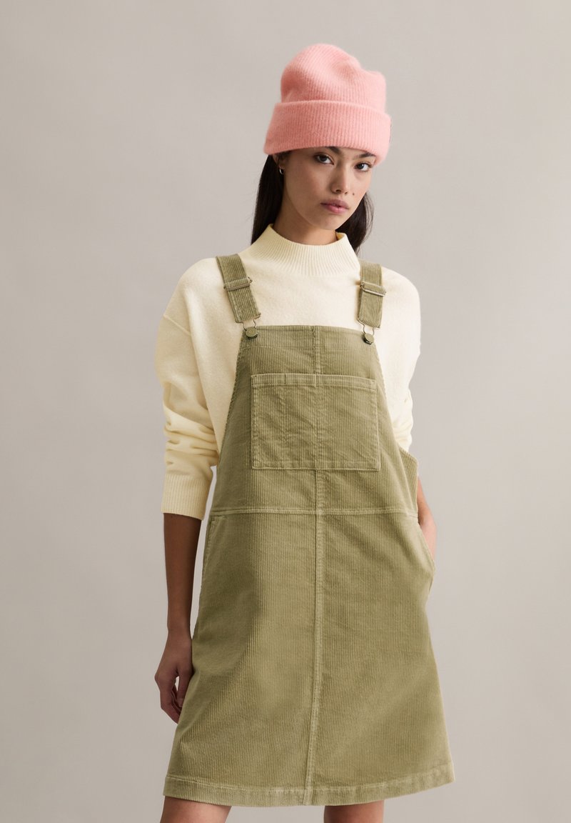 Olijfgroene corduroy salopetjurk met verstelbare bandjes, grote voorzak, gedragen over een crèmekleurige gebreide trui, afgewerkt met een roze beanie.