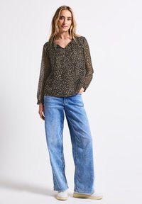 Camicia trasparente a maniche lunghe in verde oliva scuro con stampa leopardata nera, abbinata a jeans in denim azzurri a gamba larga e sneakers bianche.