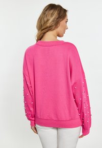 Sudadera rosa de gran tamaño con mangas largas adornada con embellishments en plata y blanco. Presenta un cuello y puños y dobladillo acanalados.