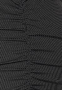 Il tessuto nero a coste presenta una texture verticale con cuciture arricciate, creando una silhouette aderente. Nessuna hardware o motivo visibile.