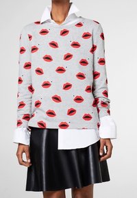 Grå sweatshirt med et gentaget print af røde læber foran, parret med en hvid skjorte med knapper under, og en sort lædernederdel.