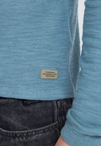 Chemise à manches longues texturée bleu clair avec une étiquette cousue affichant du texte. Portée avec un jean en denim foncé. Design simple et décontracté.
