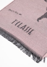 Sciarpa intrecciata in tessuto rosa tenue, con testo stampato nero. I bordi sono rifiniti con frange nere, che aggiungono texture e dettaglio al design.