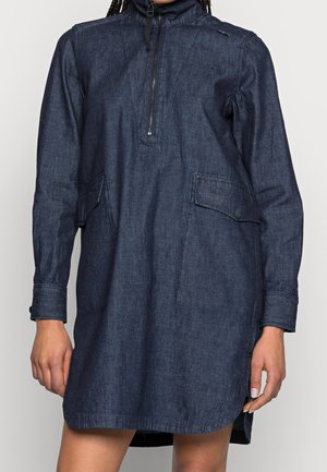 Długa sukienka z niebieskiego dżinsu z długim rękawem, z przodu z zamkiem błyskawicznym, wysokim kołnierzem i dużymi kieszeniami z patkami, noszona przez osobę z widocznymi rękami.