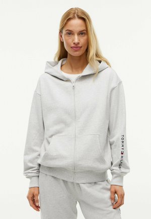 Femme portant un sweat à capuche gris clair zippé avec poches avant et pantalon de jogging assorti, debout devant un fond blanc uni.