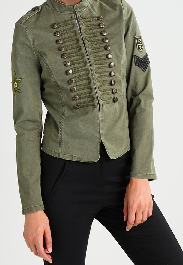 Veste militaire vert olive avec boutons décoratifs, écussons étoile et chevrons aux manches, portée avec un pantalon noir.