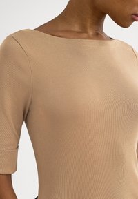 Μπεζ εφαρμοστό τοπ με λαιμόκοψη βάρκα, τρία τέταρτα μανίκια και λεία υφή. Το ύφασμα φαίνεται ελαστικό και διαπνέον.