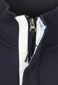 Marine blauwe ritsjack met een ribbelkraag, witte knoopsluiting en een zachte textuur. Inclusief een gegroefde rits en gestreepte binnenvoering.