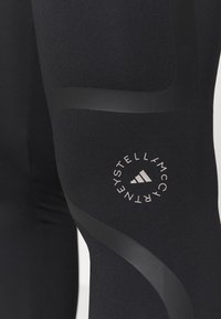 Czarne legginsy sportowe z odblaskowymi zakrzywionymi paskami i okrągłym logo Stella McCartney Adidas na udzie.