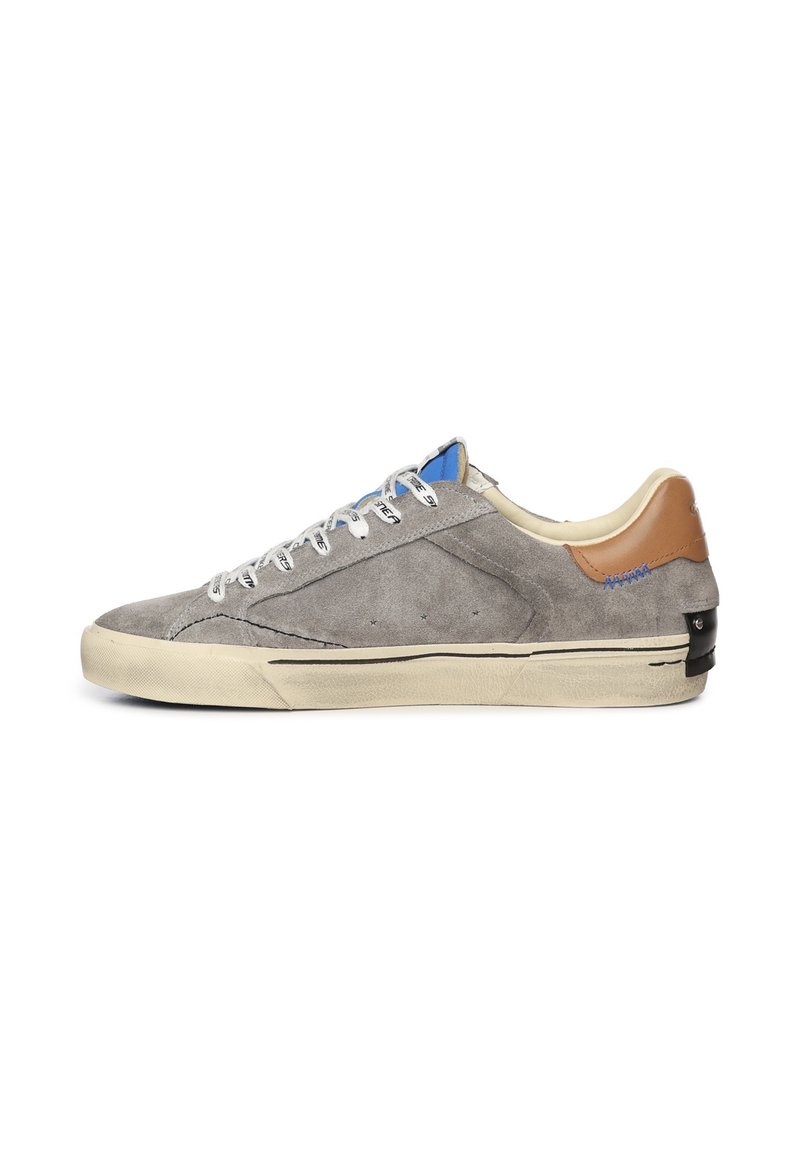 Sneaker grigio in suede con lacci bianchi, accento in pelle avana sul tallone, dettagli blu e neri, e suola in gomma testurizzata. Design minimalista.