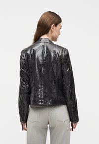 Γυναίκα γυρισμένη από πίσω, που φορά μια λαμπερή μαύρη ζακέτα με μοτίβο δέρματος φιδιού και ανοιχτογκρι τζιν, σε λευκό φόντο.