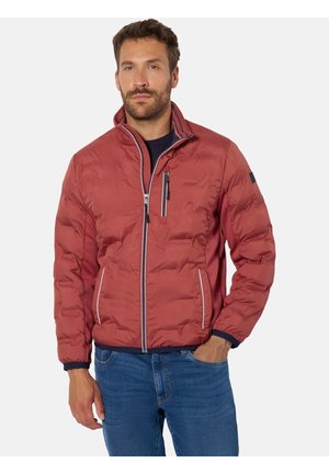 Mann trägt eine rote gesteppte Jacke mit Reißverschlusstaschen und blaue Jeans, steht mit einer Hand in der Tasche vor einem schlichten Hintergrund.