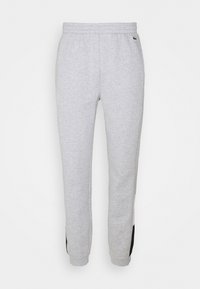 Světlé šedé tepláky s elastickým pasem, černými lemy na koncích nohavic a malým zeleným krokodýlím logem na pravém boku.