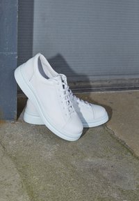 Baskets en cuir blanc avec un bout arrondi, des lacets plats et une semelle en caoutchouc blanche texturée, positionnées sur un fond gris.