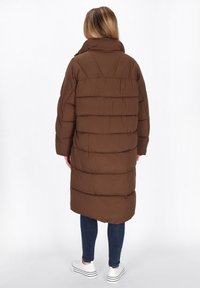 Bruine pufferjas met een hoge kraag, voorzien van horizontaal gestikte secties. Gegoed over donkere jeans en witte schoenen, met een relaxed fit.