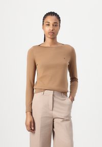 Maglione fitted marrone con maniche lunghe e scollo ampio, caratterizzato da una sottile fascia a coste e un piccolo logo sul davanti. Abbinato a pantaloni beige chiaro.