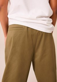 Lässig geschnittene, olivgrüne Hose aus Baumwolle, mit einer Gesäßtasche und weicher Textur, kombiniert mit einem weißen, kurzärmeligen Hemd.