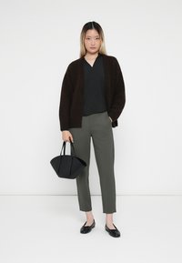 Brun strikket cardigan, svart v-hals topp, olivengrønne ankelbukser, svarte leiligheter og en svart origami-stil håndveske. Enkelt bakgrunn.