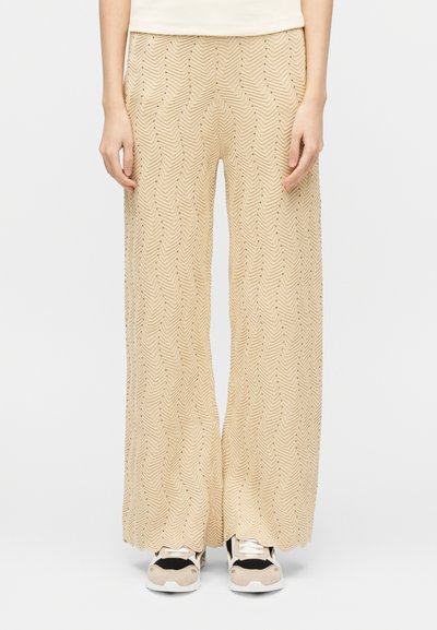Personne portant un pantalon large texturé beige et des baskets noires et blanches, debout sur un fond blanc.